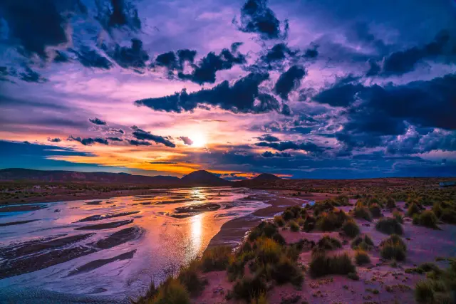 A vivid purple and orange sunset reflected in a braided river winding through scrubland.